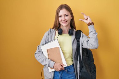 Young caucasian woman wearing student backpack and holding books smiling and confident gesturing with hand doing small size sign with fingers looking and the camera. measure concept. 