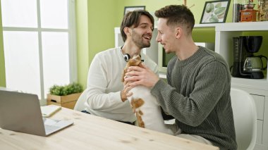 İki erkek yemek odasında köpekle aynı masada dizüstü bilgisayar kullanıyor.