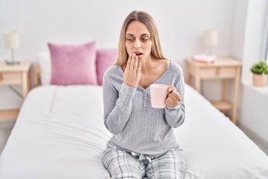 Young woman tired drinking cup of coffee sitting on bed at bedroom