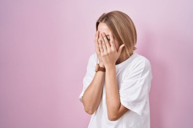 Young caucasian woman standing over pink background with sad expression covering face with hands while crying. depression concept. 