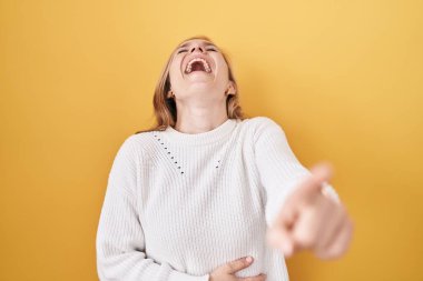 Beyaz süveter giyen, sarı arka planda beyaz bir kadın sana gülüyor, eli vücudunun üzerinde kamerayı işaret ediyor, yüz ifadesi utanç verici. 