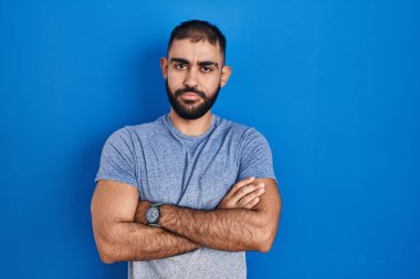 Mavi arka planda duran sakallı, şüpheci ve gergin Orta Doğulu bir adam. Yüzünde çapraz kollu tasvip etmeyen bir ifade var. negatif kişi. 