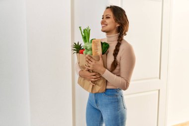 Genç, güzel, İspanyol bir kadın gülümsüyor. Elinde alışveriş torbası var.