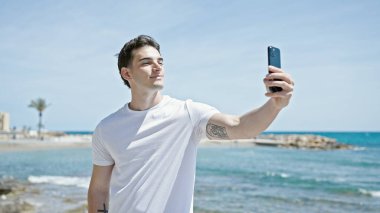 Genç İspanyol adam gülümsüyor sahildeki akıllı telefonun yanında özçekim yapıyor.
