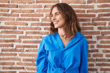 Güzel esmer kadın tuğla duvarın üzerinde duruyor. Yüzünde gülümseme, doğal bir ifade ile yan yana bakıyor. Kendinden emin gülen. 