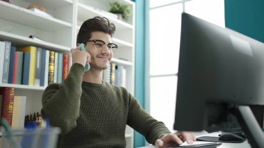 Young hispanic man student talking on smartphone using computer at library university