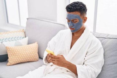 Young latin man relaxed on sofa with facial mask treatment using smartphone at home