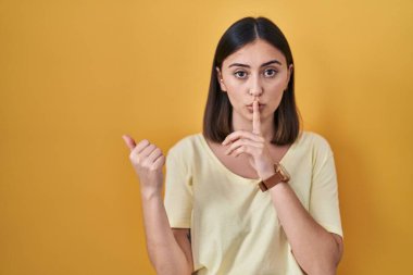 Sarı arka plan üzerine gündelik tişört giyen İspanyol kız sessiz olmak istiyor. Parmakları dudaklarında, elleri yana doğru işaret ediyor. sessizlik ve gizli konsept. 