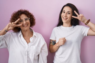İspanyol anne ve kızı pembe arka planda günlük beyaz t-shirt giyiyorlar. Barış sembolü yapıyorlar. Yüzlerinde parmaklar, gülümseyerek zafer gösteriyorlar. 