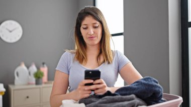 Young blonde woman using smartphone hanging clothes on clothesline at laundry room