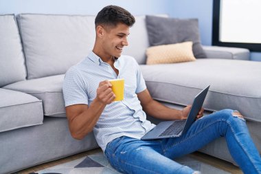 Genç İspanyol adam dizüstü bilgisayar kullanıyor ve evde oturmuş kahve içiyor.