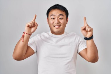 Beyaz arka planda duran genç Çinli adam şaşırmış, şaşırmış ve parmaklarıyla işaret ediyor ve kollarını kaldırıyordu.. 