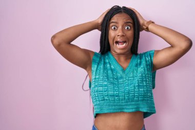 Genç Afro-Amerikan örgüleri pembe arka planda çılgın bir şekilde duruyor ve elleri başının üstünde korkmuş ve ağzı açık bir şekilde şok geçirmekten korkuyor. 