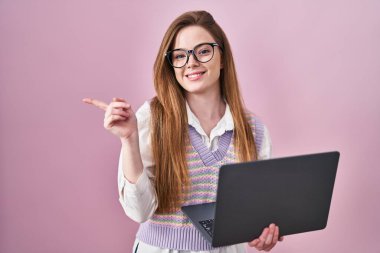 Yüzünde kocaman bir gülümseme olan bilgisayar bilgisayarı kullanan beyaz bir kadın kameraya el ile işaret ediyor.. 