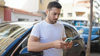 Young hispanic man using smartphone leaning on car at street