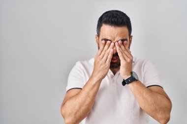 Sakallı, beyaz arka plan üzerine günlük kıyafetler giyen, yorgun ve yorgun görünen, uykulu ve yorgun ifadeli İspanyol bir genç. görme sorunu 