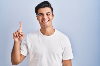 Mavi arka planda duran İspanyol adam bir numarayı gösterip işaret ederken kendinden emin ve mutlu gülümsüyor.. 