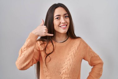 Genç esmer kadın beyaz arka planda durup gülümsüyor telefonla el ve parmaklarıyla telefon hareketi yapıyor telefonda konuşuyor gibi. iletişim kavramları. 