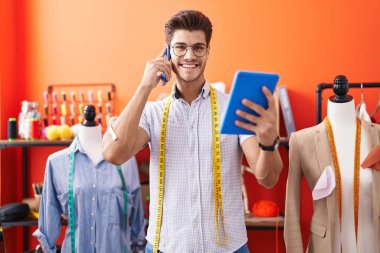 Young hispanic man tailor talking on smartphone using touchpad at atelier