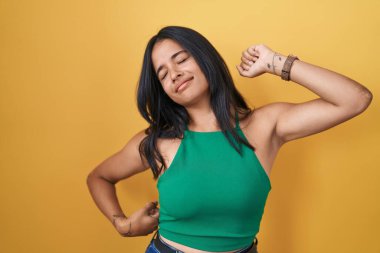 Esmer kadın sarı arka planda duruyor, geriniyor, yorgun ve rahat, uykulu ve sabahın erken saatleri için esniyor. 