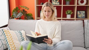 Young blonde woman reading book sitting on sofa at home