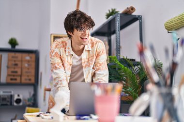 Young hispanic man artist smiling confident using laptop at art studio