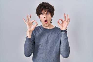 Young man standing over isolated background looking surprised and shocked doing ok approval symbol with fingers. crazy expression 