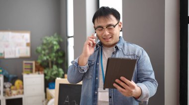 Young chinese man preschool teacher using touchpad talking on smartphone at kindergarten
