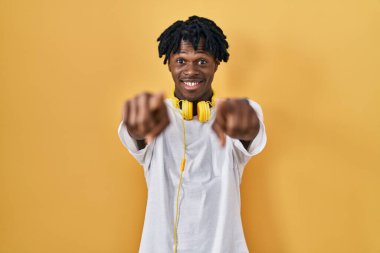 Sarı arka planda duran Afrikalı genç adam parmaklarıyla seni ve kamerayı işaret ediyor, neşeli ve pozitif gülümsüyor. 