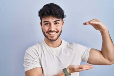 Hispanic man with beard standing over white background gesturing with hands showing big and large size sign, measure symbol. smiling looking at the camera. measuring concept. 