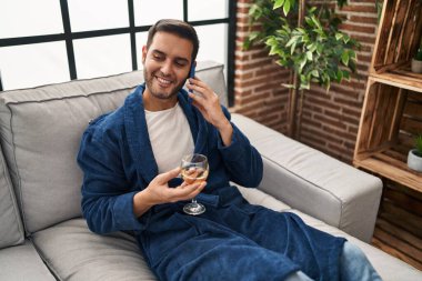 Young hispanic man talking on the smartphone and drinking wine at home