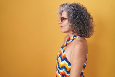 Sarı arka planda duran gri saçlı orta yaşlı bir kadın. Yan tarafa bakıyor, rahat bir görünüm takınıyor ve kendine güvenen bir gülüşü var.. 