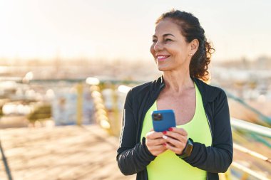Middle age hispanic woman working out with smartphone at promenade