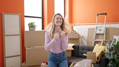 Genç sarışın kadın kendine güvenerek gülümsüyor. Kazanan yüz ifadesiyle yeni evinde duruyor.