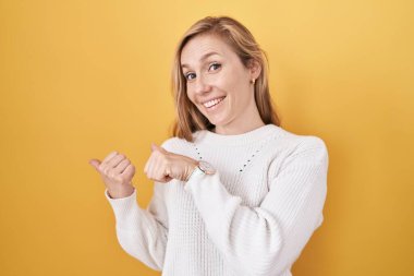 Sarı arka planda beyaz süveter giyen genç, beyaz bir kadın. Arka tarafı işaret ediyor. El ve baş parmakları havada, kendinden emin gülümsüyor. 