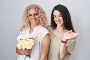 Anne ve kızı ellerinde bir buket beyaz çiçek tutarak mutlu bir şekilde sunum yapıyor ve avuç içiyle kameraya bakıyor.. 