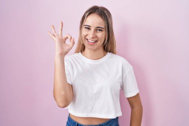 Young blonde woman standing over pink background smiling positive doing ok sign with hand and fingers. successful expression. 