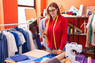 Young beautiful plus size woman tailor smiling confident holding scissors at atelier