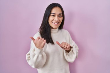 Pembe arka planda duran genç Güney Asyalı kadın arka tarafı işaret ediyor, el ve baş parmakları havada, kendinden emin gülümsüyor. 