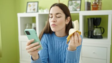 Beyaz kadın yemek odasında akıllı telefon kullanarak muz yiyor.