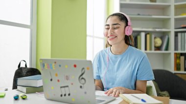 Genç ve güzel İspanyol öğrenci kütüphanede bilgisayar ve kulaklık kullanıyor.
