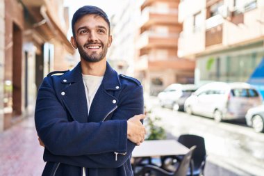 Kahve dükkanının terasında kolları çapraz hareketlerle duran İspanyol genç adam.
