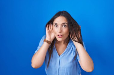 Young brunette woman standing over blue background trying to hear both hands on ear gesture, curious for gossip. hearing problem, deaf 