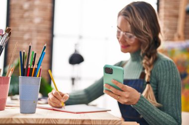 Sanat stüdyosunda akıllı telefon kullanarak not defterine çizim yapan genç bir kadın sanatçı.