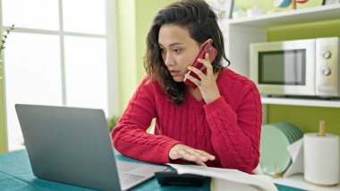 Genç, güzel, İspanyol bir kadın yemek odasında bilgisayarla akıllı telefondan konuşuyor.