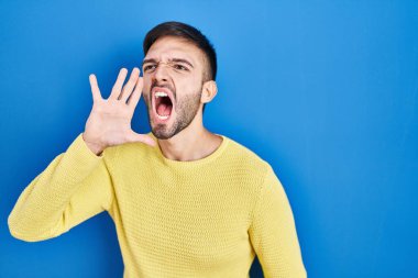 Hispanic man standing over blue background shouting and screaming loud to side with hand on mouth. communication concept. 
