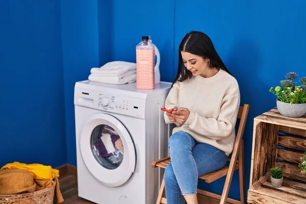 年轻美丽的惊慌失措的女人用智能手机在洗衣房等洗衣机 — 图库照片