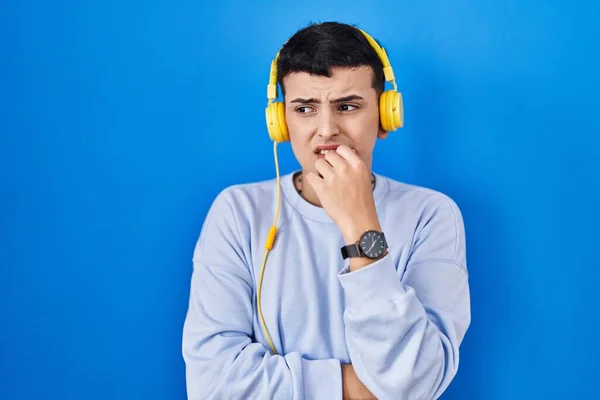 Non binary person listening to music using headphones looking stressed and nervous with hands on mouth biting nails. anxiety problem.