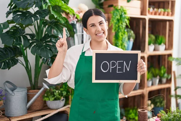 Young hispanic woman working at florist holding open sign smiling with an idea or question pointing finger with happy face, number one