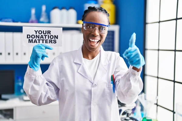 Beautiful black woman working at scientist laboratory holding your donation matters banner smiling happy and positive, thumb up doing excellent and approval sign